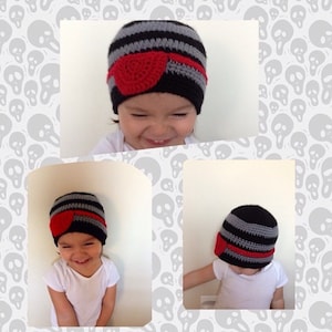 Pode incluir: Uma criança usando um chapéu de tricô listrado preto e cinza com um tapa-olho de crochê vermelho. O chapéu tem um padrão de listras pretas e cinzas.