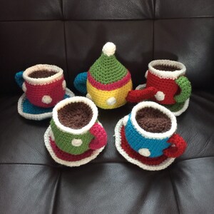 Op de afbeelding: Een gehaakt theeservies met een gele theepot, groene, rode en blauwe theekopjes en witte schoteltjes. De theekopjes hebben witte stippen.