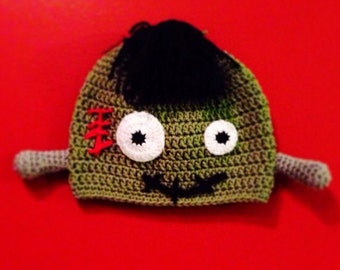 May include: Green crocheted hat with black yarn hair, white eyes, and a red stitched scar. The hat has a black yarn moustache and two small green yarn arms.