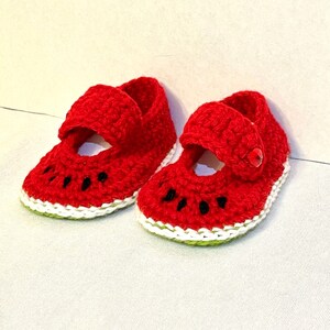 Puede incluir: Un par de zapatos de beb&eacute; de ganchillo rojo con forma de rodajas de sand&iacute;a. Los zapatos tienen ribetes blancos y verdes y costuras negras para las semillas.