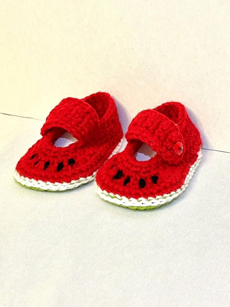 Puede incluir: Un par de zapatos de beb&eacute; de ganchillo rojo con forma de rodajas de sand&iacute;a. Los zapatos tienen ribetes blancos y verdes y costuras negras para las semillas.