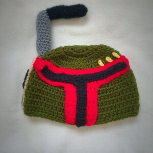 May include: Green, red, and black crocheted hat shaped like a Boba Fett helmet. The hat has a grey and black crocheted antenna on top.