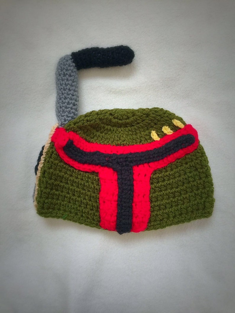 May include: Green, red, and black crocheted hat shaped like a Boba Fett helmet. The hat has a grey and black crocheted antenna on top.