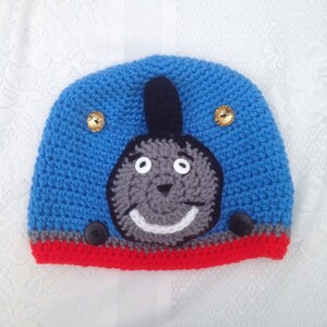 May include: Blue and red crocheted hat with a grey Thomas the Tank Engine face on the front. The hat has two gold buttons on the top and one black button on the side.