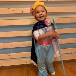 May include: A young child dressed as a character from the movie Toy Story. They are wearing a blue and pink dress with a yellow and pink hat. They are holding a blue wand and smiling at the camera.