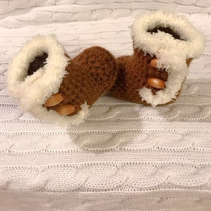 May include: A pair of brown crocheted baby booties with white faux fur trim and brown wooden buttons.
