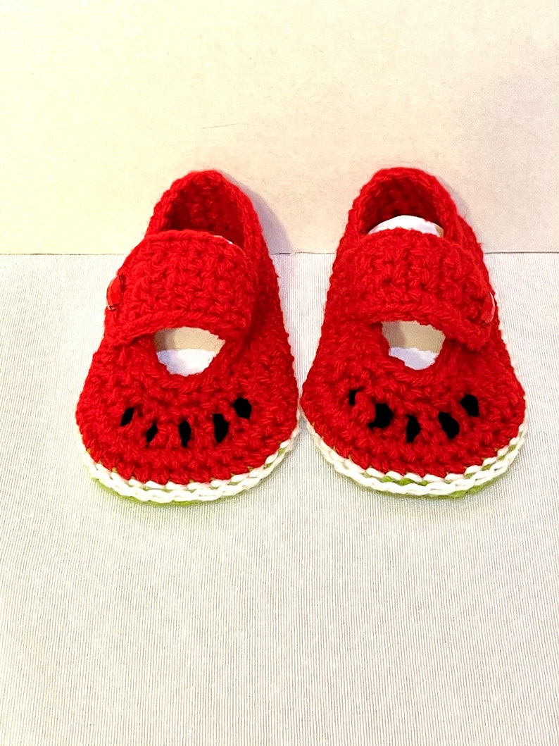 Puede incluir: Un par de zapatos de beb&eacute; de ganchillo rojo con forma de rodajas de sand&iacute;a. Los zapatos tienen un borde blanco y semillas negras.