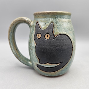 May include: A ceramic mug with a speckled light blue and green glaze featuring a raised, matte black cat silhouette on the front. The cat has two wide, light-colored eyes and a long tail that curves along the bottom of the mug's rounded body.