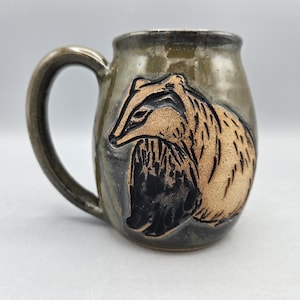 May include: A ceramic mug with a glossy, dark olive-green glaze and a large, curved handle. The front features a carved, unglazed tan-colored badger illustration with black ink detailing, showcasing the animal in a seated profile position.