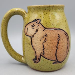 May include: A ceramic mug with a light green speckled glaze and a brown capybara illustration. The mug has a large handle and a cylindrical shape, ideal for tea or coffee.