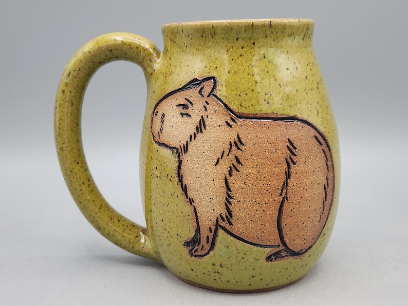 May include: A ceramic mug with a light green speckled glaze and a brown capybara illustration. The mug has a large handle and a cylindrical shape, ideal for tea or coffee.