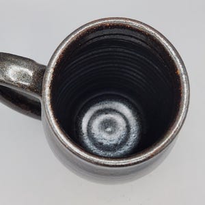 Puede incluir: Taza de cer&aacute;mica hecha a mano con exterior marr&oacute;n oscuro y borde marr&oacute;n claro contrastante. El interior es negro profundo y brillante. La taza tiene forma redondeada y un asa robusta.