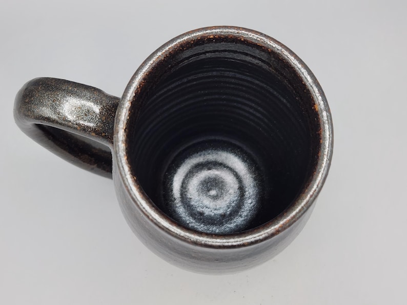 Puede incluir: Taza de cer&aacute;mica hecha a mano con exterior marr&oacute;n oscuro y borde marr&oacute;n claro contrastante. El interior es negro profundo y brillante. La taza tiene forma redondeada y un asa robusta.