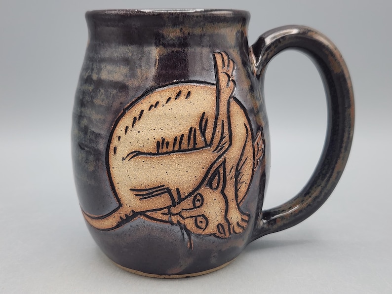 Może przedstawiać: Ciemny, błyszczący ceramiczny kubek z ręcznie wyrytym jasnym wzorem zwiniętego w kłębek kota. Kot jest przedstawiony z podniesioną tylną łapą, a kubek posiada solidne ucho oraz gładki, zaokrąglony kształt korpusu.