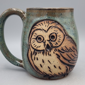 May include: Handmade ceramic mug with a detailed owl design. The mug features a brown owl illustration against a teal and brown background. The handle and rim have a speckled, earthy appearance, adding to its rustic charm.