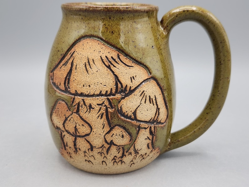 Puede incluir: Una taza de cer&aacute;mica hecha a mano con un esmalte verde bosque. La taza presenta un dise&ntilde;o en relieve detallado de varios hongos en tonos marrones y beige. El asa es curvada y la taza tiene una est&eacute;tica r&uacute;stica y terrosa.