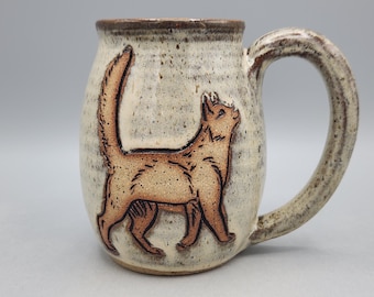 May include: A ceramic mug with a speckled off-white glaze and a dark brown rim. The mug features a raised, embossed illustration of a cat in a light brown tone with dark brown outlines, depicted in a walking pose with its tail held high.