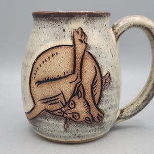 Może przedstawiać: Ceramiczny kubek z nakrapianym, kremowobiałym szkliwem i brązowym rantem. Z przodu znajduje się wyryta, beżowa ilustracja kota myjącego się z uniesioną łapą. Uchwyt jest zakrzywiony i pasuje do nakrapianego wykończenia całego naczynia.