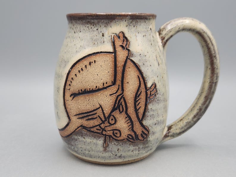 Może przedstawiać: Ceramiczny kubek z nakrapianym, kremowobiałym szkliwem i brązowym rantem. Z przodu znajduje się wyryta, beżowa ilustracja kota myjącego się z uniesioną łapą. Uchwyt jest zakrzywiony i pasuje do nakrapianego wykończenia całego naczynia.