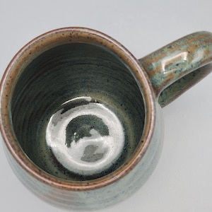 May include: A handmade ceramic mug with a rounded body and a large handle. The mug features a speckled, earthy colour palette with shades of green, brown, and tan. The interior is a darker green, and the rim has a contrasting brown band.