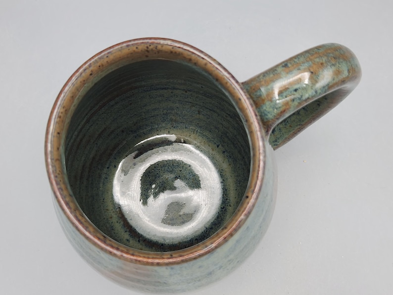 May include: A handmade ceramic mug with a rounded body and a large handle. The mug features a speckled, earthy colour palette with shades of green, brown, and tan. The interior is a darker green, and the rim has a contrasting brown band.