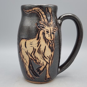 Puede incluir: Taza de cer&aacute;mica hecha a mano con un esmalte gris oscuro y una ilustraci&oacute;n de cabra marr&oacute;n. La taza tiene un asa curva y una forma cil&iacute;ndrica. El dise&ntilde;o de la cabra est&aacute; grabado en la superficie, creando un efecto texturizado. Para bebidas fr&iacute;as o calientes.