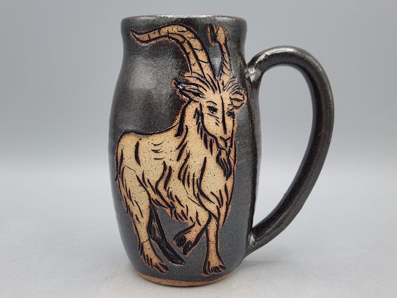 Puede incluir: Taza de cer&aacute;mica hecha a mano con un esmalte gris oscuro y una ilustraci&oacute;n de cabra marr&oacute;n. La taza tiene un asa curva y una forma cil&iacute;ndrica. El dise&ntilde;o de la cabra est&aacute; grabado en la superficie, creando un efecto texturizado. Para bebidas fr&iacute;as o calientes.