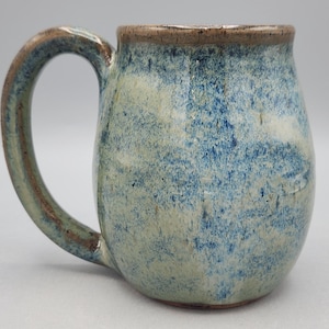 Może przedstawiać: Ceramiczny kubek z nakrapianą niebiesko-kremową glazurą, zaokrąglonym korpusem i zakrzywionym uchwytem. Krawędź i brzegi uchwytu są podkreślone brązowawym, ziemistym odcieniem, co nadaje całej powierzchni teksturalny wygląd.