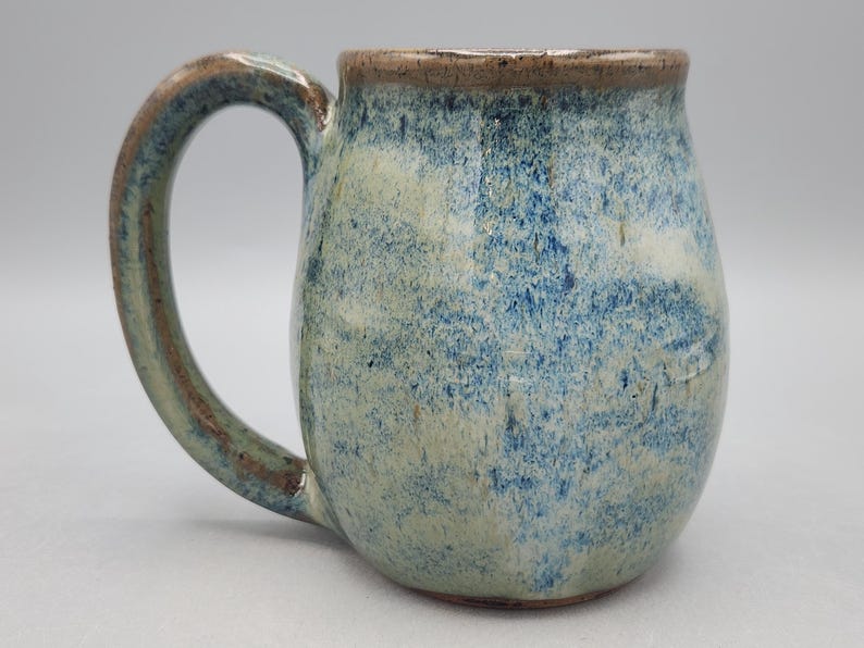 Może przedstawiać: Ceramiczny kubek z nakrapianą niebiesko-kremową glazurą, zaokrąglonym korpusem i zakrzywionym uchwytem. Krawędź i brzegi uchwytu są podkreślone brązowawym, ziemistym odcieniem, co nadaje całej powierzchni teksturalny wygląd.