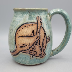Może przedstawiać: Ceramiczny kubek z jasnoniebiesko-zielonym nakrapianym szkliwem, ozdobiony ciemnobrązową, wygrawerowaną ilustracją kota myjącego się. Kubek ma zaokrąglony korpus, szeroki brzeg oraz solidne, zakrzywione ucho po prawej stronie naczynia.