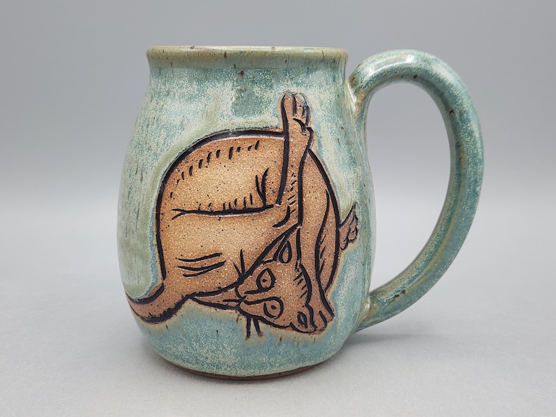 Może przedstawiać: Ceramiczny kubek z jasnoniebiesko-zielonym nakrapianym szkliwem, ozdobiony ciemnobrązową, wygrawerowaną ilustracją kota myjącego się. Kubek ma zaokrąglony korpus, szeroki brzeg oraz solidne, zakrzywione ucho po prawej stronie naczynia.