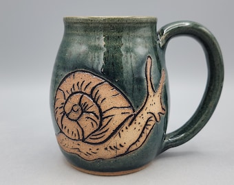 May include: A ceramic mug with a dark green glazed finish and a large, curved handle. The front of the mug features a detailed, embossed illustration of a snail with a textured, light brown shell and body, contrasting against the dark glaze.