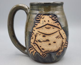 May include: A ceramic mug with a glossy, dark, mottled green and brown glaze. The front features a raised, unglazed, light tan relief of a seated toad with detailed etched lines defining its eyes, mouth, and textured skin.