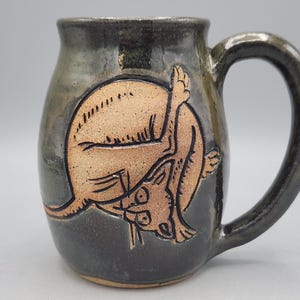 Może przedstawiać: Ciemnoszary, błyszczący kubek ceramiczny z wygrawerowaną, nieszkliwioną ilustracją w kolorze opalenizny, przedstawiającą zwiniętego w kłębek kota. Kubek ma zaokrąglony korpus, szeroki brzeg oraz solidne, zakrzywione ucho po prawej stronie.