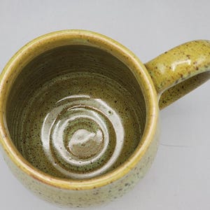 Puede incluir: Una taza de cer&aacute;mica hecha a mano con un interior verde oliva moteado y un exterior amarillo-verdoso. La taza presenta un cuerpo redondeado y un asa curvada. El interior tiene un patr&oacute;n en espiral. La taza es adecuada para bebidas fr&iacute;as o calientes.