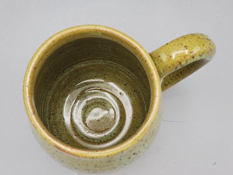 Puede incluir: Una taza de cer&aacute;mica hecha a mano con un interior verde oliva moteado y un exterior amarillo-verdoso. La taza presenta un cuerpo redondeado y un asa curvada. El interior tiene un patr&oacute;n en espiral. La taza es adecuada para bebidas fr&iacute;as o calientes.