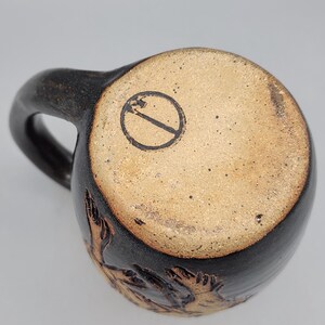 Puede incluir: Una taza de cer&aacute;mica hecha a mano con un exterior marr&oacute;n oscuro y una base marr&oacute;n claro. La taza tiene un asa curva y un sello circular en la parte inferior. La base de la taza tiene una superficie texturizada con un dise&ntilde;o tallado.