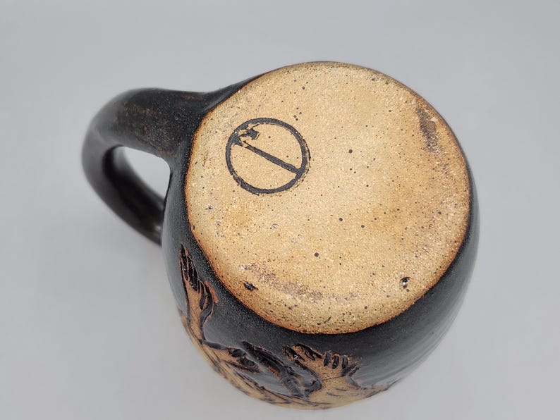 Puede incluir: Una taza de cer&aacute;mica hecha a mano con un exterior marr&oacute;n oscuro y una base marr&oacute;n claro. La taza tiene un asa curva y un sello circular en la parte inferior. La base de la taza tiene una superficie texturizada con un dise&ntilde;o tallado.