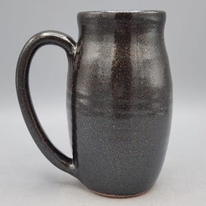 Puede incluir: Taza de cer&aacute;mica gris oscuro hecha a mano con un asa grande. La taza tiene una superficie texturizada y un borde ligeramente ensanchado. El color gris oscuro tiene sutiles reflejos met&aacute;licos, lo que le da un aspecto &uacute;nico y artesanal.