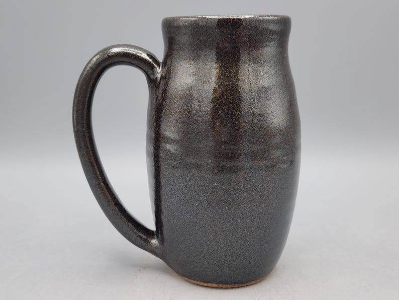 Puede incluir: Taza de cer&aacute;mica gris oscuro hecha a mano con un asa grande. La taza tiene una superficie texturizada y un borde ligeramente ensanchado. El color gris oscuro tiene sutiles reflejos met&aacute;licos, lo que le da un aspecto &uacute;nico y artesanal.