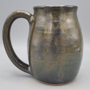 Może przedstawiać: Ceramiczny kubek o zaokrąglonym korpusie i dużym, zakrzywionym uchwycie. Powierzchnia pokryta jest nakrapianym szkliwem w odcieniach ciemnego brązu, antracytu i metalicznego brązu, co tworzy błyszczące, nier&oacute;wne wykończenie na całej powierzchni.