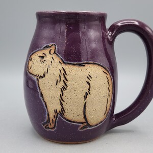 May include: A ceramic mug with a deep purple glaze and a large handle. The mug features a tan capybara illustration. The mug is a unique piece of drinkware, perfect for coffee, tea, or other beverages.