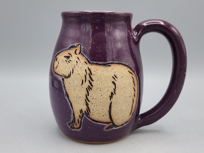 May include: A ceramic mug with a deep purple glaze and a large handle. The mug features a tan capybara illustration. The mug is a unique piece of drinkware, perfect for coffee, tea, or other beverages.