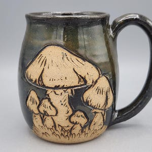 Puede incluir: Taza de cer&aacute;mica hecha a mano con un esmalte oscuro y moteado. La taza presenta un dise&ntilde;o en relieve de varios hongos en tonos beige y marr&oacute;n. El asa es de color oscuro.