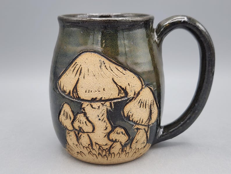 Puede incluir: Taza de cer&aacute;mica hecha a mano con un esmalte oscuro y moteado. La taza presenta un dise&ntilde;o en relieve de varios hongos en tonos beige y marr&oacute;n. El asa es de color oscuro.