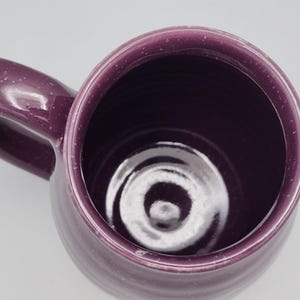 May include: A handmade, purple ceramic mug with a rounded body and a large handle. The mug has a speckled finish and a dark interior. The mug is empty and sits on a white surface.