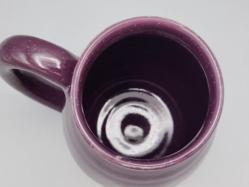 May include: A handmade, purple ceramic mug with a rounded body and a large handle. The mug has a speckled finish and a dark interior. The mug is empty and sits on a white surface.