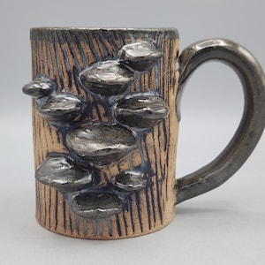Może przedstawiać: Ceramiczny kubek o teksturowanej beżowej powierzchni z pionowymi ciemnobrązowymi liniami i kilkoma wypukłymi, ciemnymi metaliczno-szarymi kształtami grzybów na zewnątrz. Kubek posiada zakrzywione ucho w pasującym ciemnym metalicznym wykończeniu.