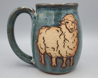 May include: A ceramic mug with a speckled blue and green glaze featuring a raised, unglazed tan relief of a sheep on the front. The mug has a curved handle and a smooth, rounded rim, showcasing a handcrafted texture and earthy color palette.