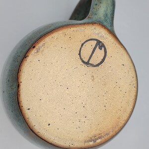 May include: A handmade ceramic mug with a rounded body and a handle. The mug has a speckled, light brown base and a teal-coloured exterior with a darker rim. A black circular stamp is visible on the base.